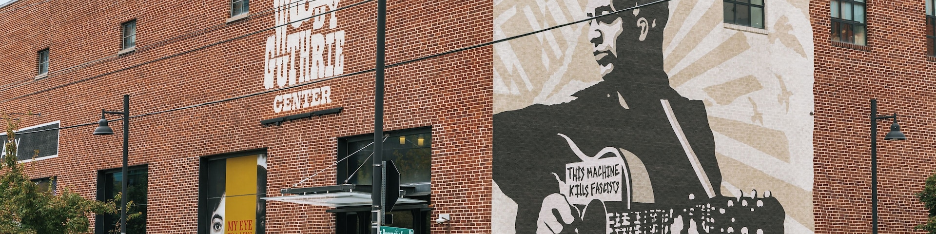 Edifício de tijolos com um mural de um homem segurando um violão e uma placa com a inscrição 'THIS MACHINE KILLS FASCISTS'.