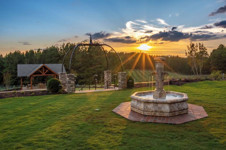 A gazebo, a stone fountain, and a sunset over a grassy field.