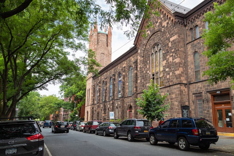 A historic church with tall spires and large windows, surrounded by trees and parked cars on a city street.