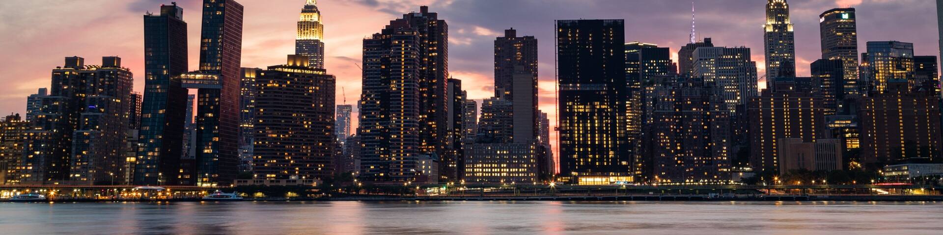 A city skyline at dusk with prominent buildings including a tall skyscraper with a spire.