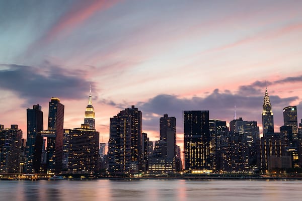 Une vue sur les toits d’une ville au crépuscule, avec des bâtiments imposants, dont un gratte-ciel doté d’une flèche.