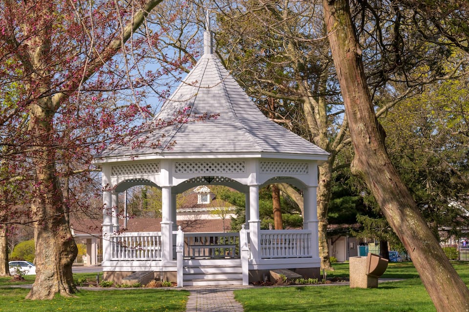 A gazebo in a park with a paved path leading to it, surrounded by trees and grass.