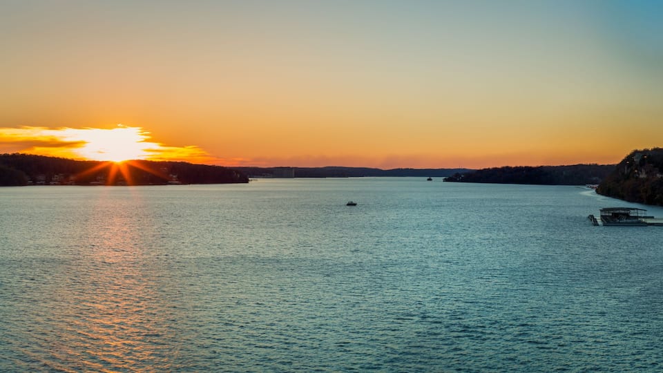 A sunset over a calm lake with boats, a rocky shoreline, and a forested hill in the background.