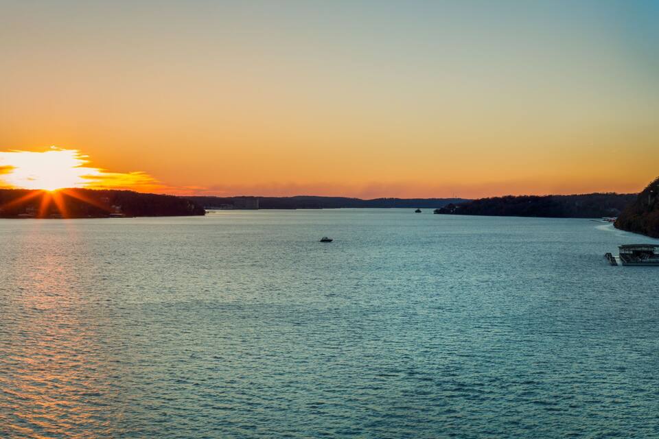 A sunset over a calm lake with boats, a rocky shoreline, and a forested hill in the background.