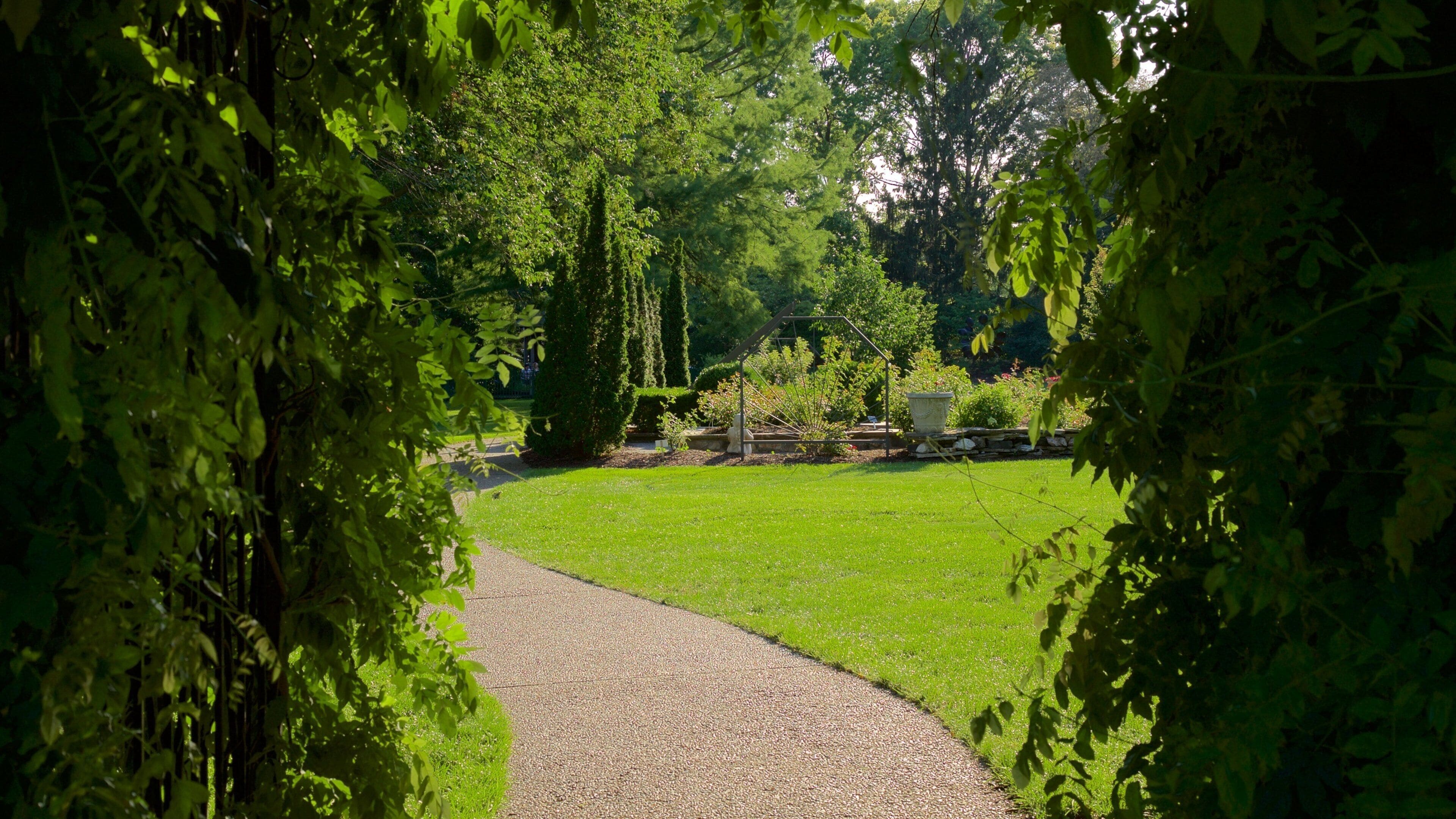 Un sendero de grava serpentea por un exuberante jardín con césped verde, árboles y un muro de piedra.