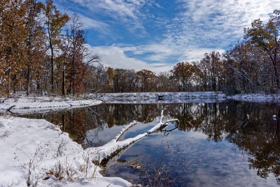 A snow-covered landscape with a calm lake reflecting trees and sky.