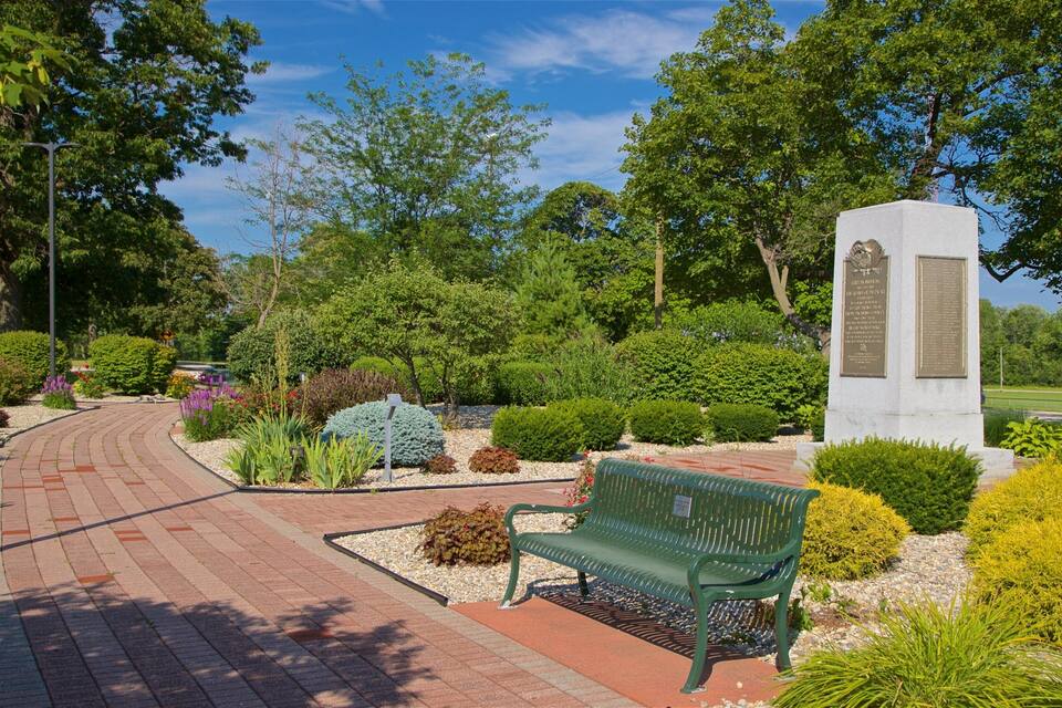 Un parque con un camino pavimentado, un banco verde y un monumento con una inscripción.