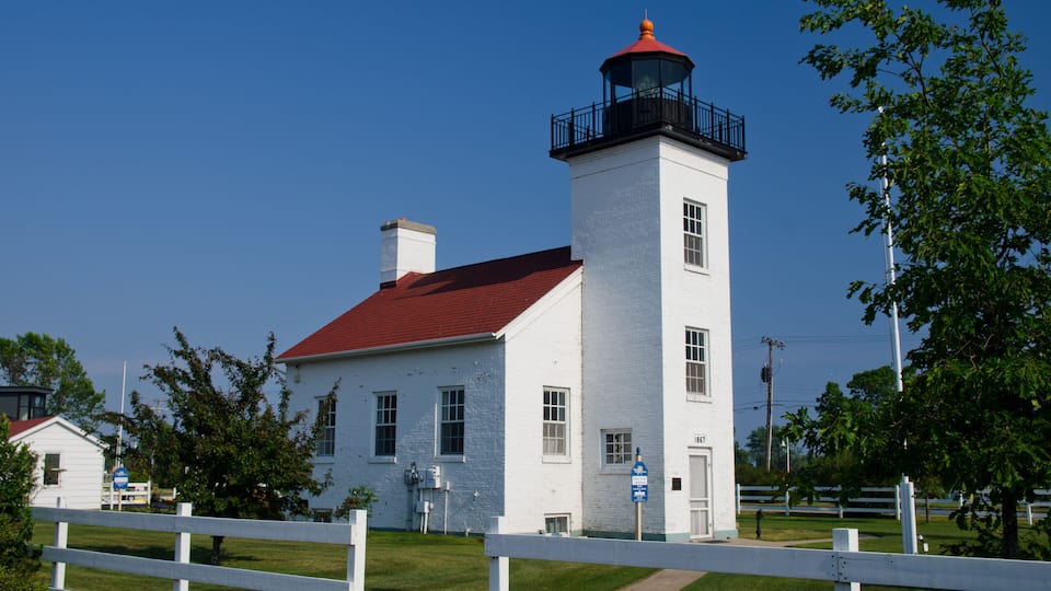 A white lighthouse with a red roof and a black balcony, surrounded by a white fence and greenery.