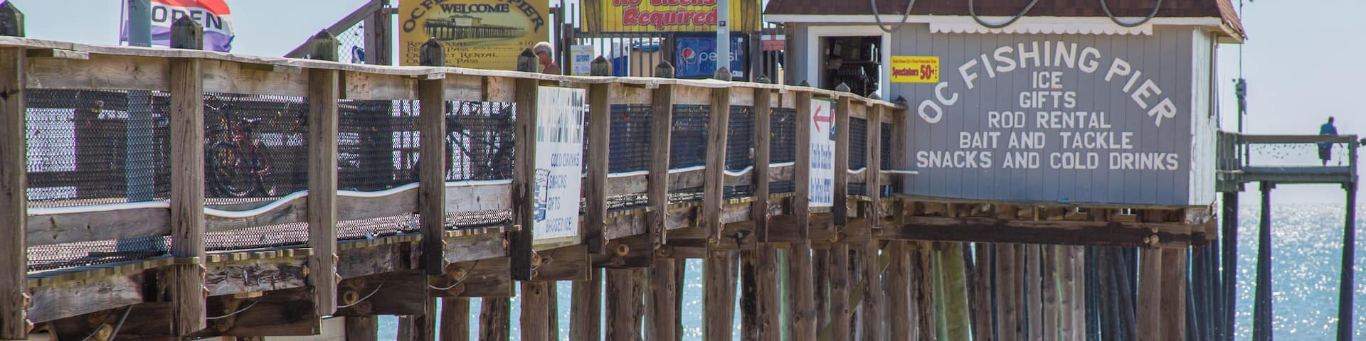 Un muelle de madera con un cartel que anuncia servicios para pescadores, incluyendo hielo, regalos, alquiler de cañas, cebo, aparejos, aperitivos y bebidas frías.