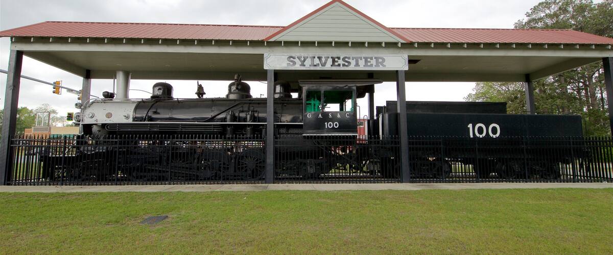 A vintage steam locomotive at a station named Sylvester, with a covered waiting area and a number 100 sign.