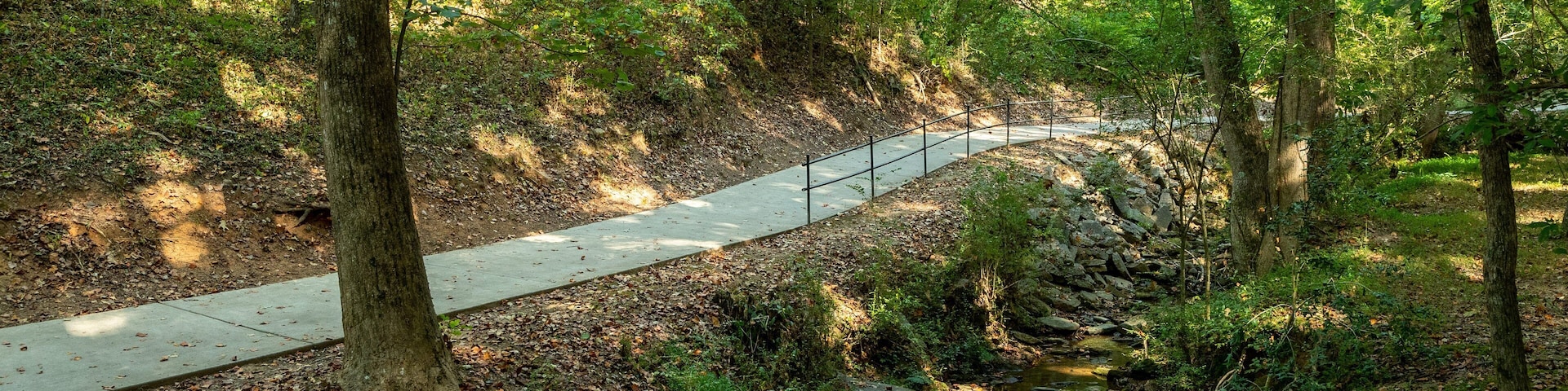 Uma área arborizada com um caminho de concreto, um pequeno riacho e uma cerca de metal.