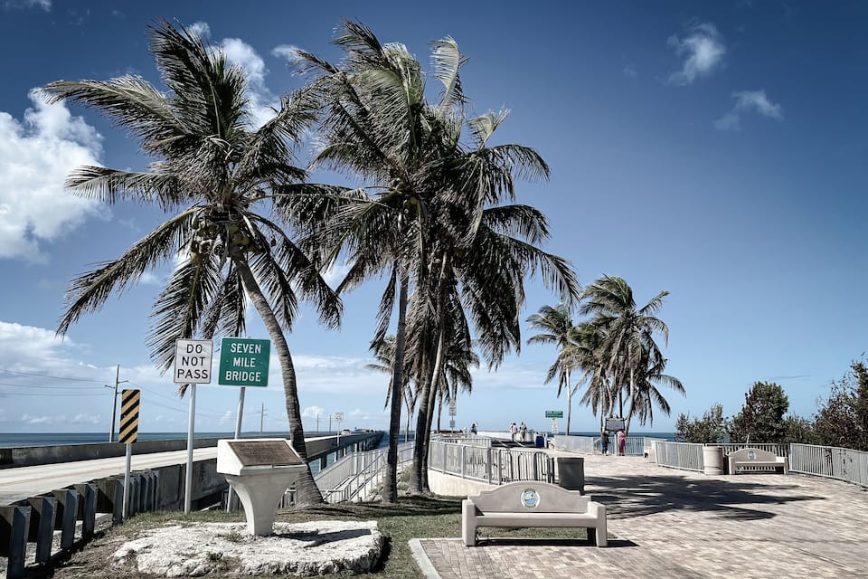 Una playa con palmeras, un letrero de 'NO PASAR', un letrero que dice 'SEVEN MILE BRIDGE' y un banco.