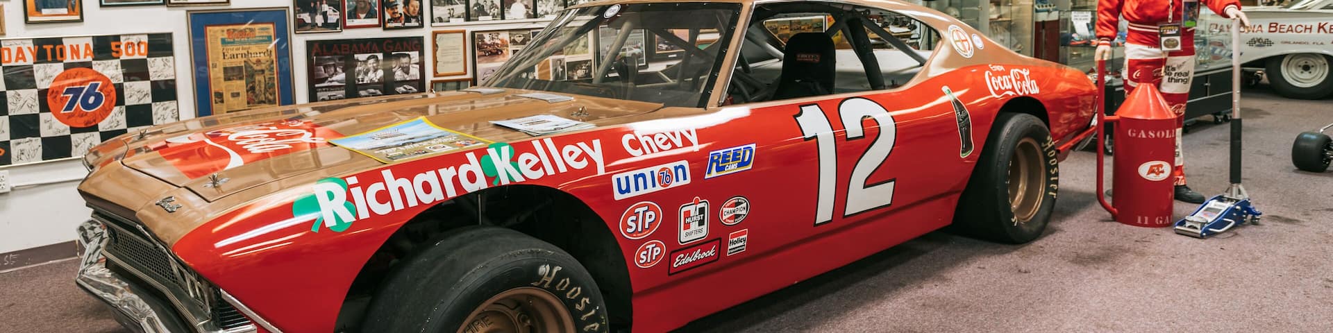 A vintage race car with the number 12, adorned with various sponsor logos, parked in a garage filled with memorabilia.