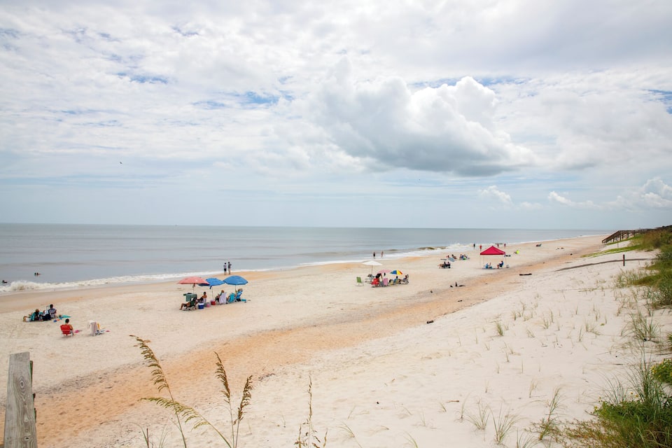 A sandy beach with people relaxing under umbrellas, a wooden fence, and dunes in the foreground.