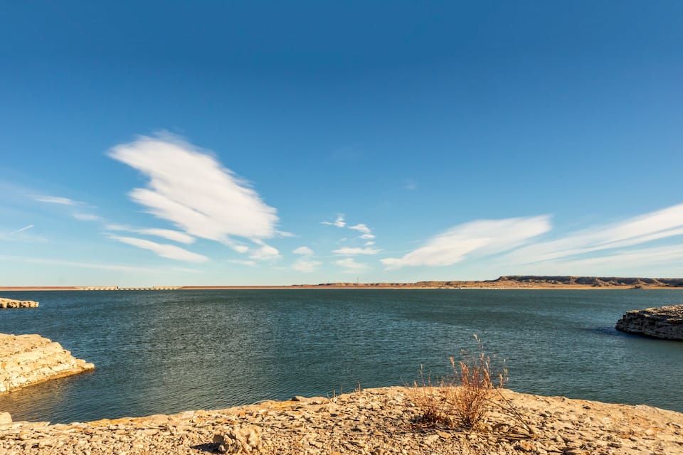 Helderblauwe lucht, kalm water, een rotsachtige kustlijn en land in de verte.