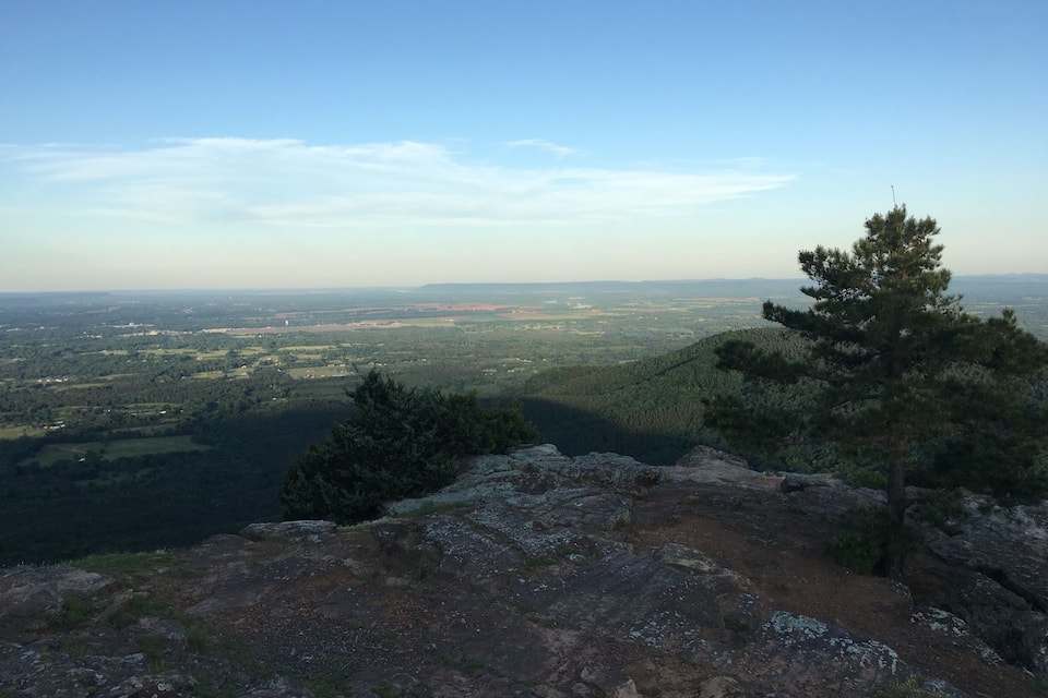 A rocky mountain ridge with a pine tree and a view of a valley with fields and distant hills.