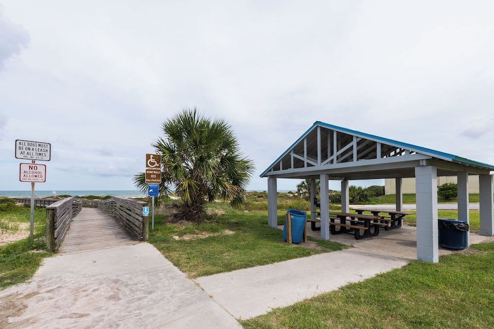 A beachside walkway leading to a covered picnic area with benches and a trash bin.