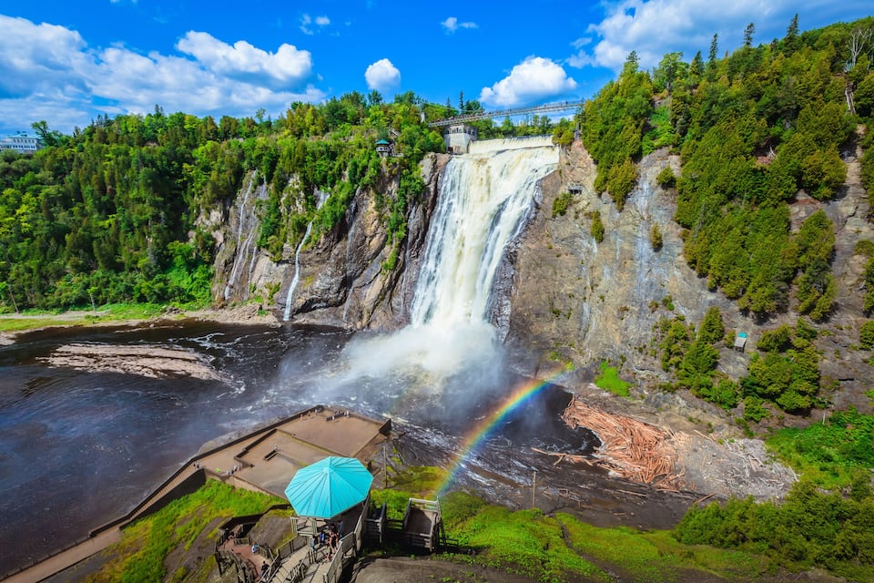 A waterfall, a rainbow, and a forested area with a bridge in the background.