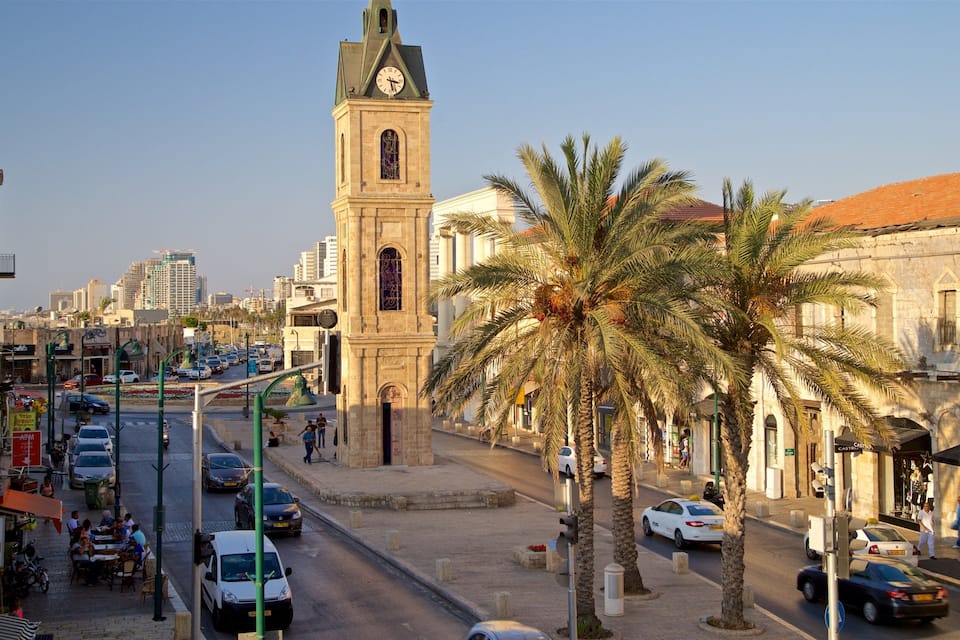 Uma rua urbana com uma torre do relógio imponente, palmeiras e diversos edifícios.