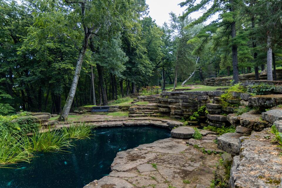 Un bosque tranquilo con un estanque azul cristalino, rodeado de escalones de piedra y abundante vegetación.