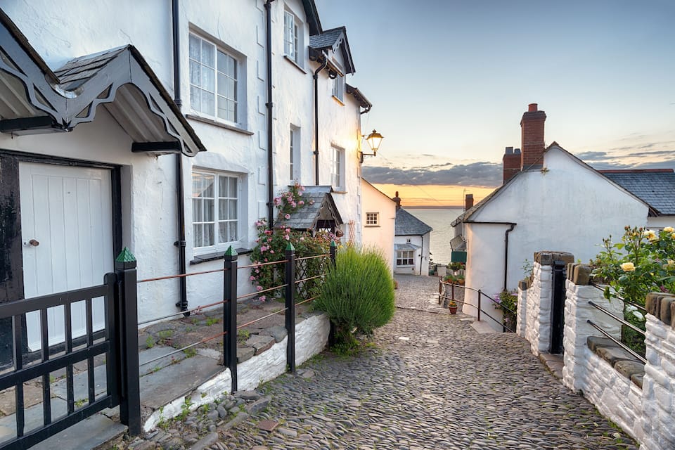 A cobblestone street in a historic town with white buildings, a black gate, and a sunset in the background.