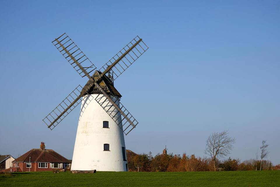 Eine weiße Windmühle mit Holzkonstruktion vor blauem Himmel, im Hintergrund ein Haus und Bäume.