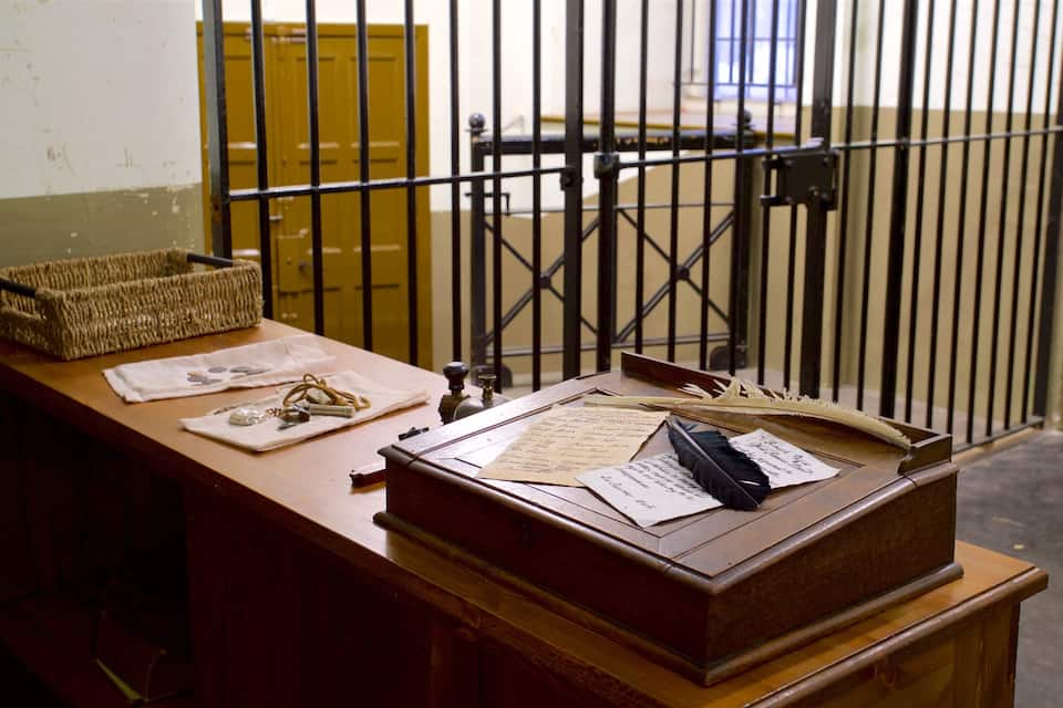 A wooden desk with papers, a quill, and a basket in a room with barred windows.