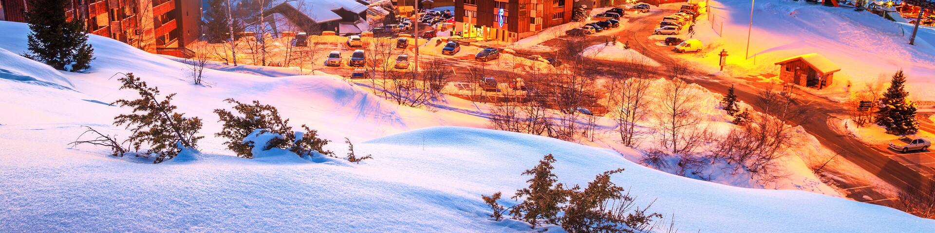 A snow-covered mountain town at sunset with buildings and a ski slope.