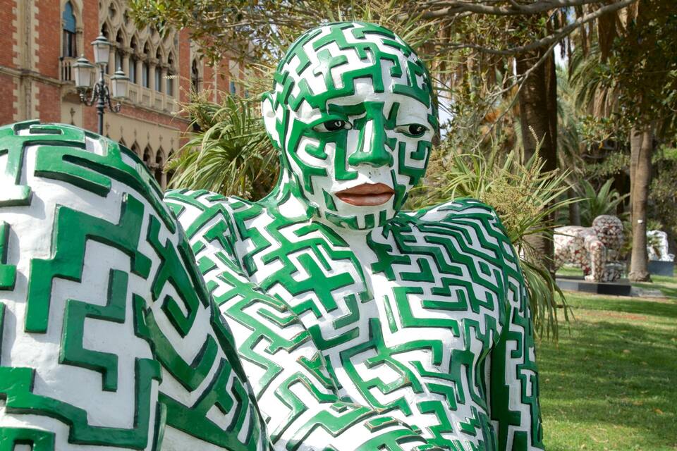 Uma pessoa com um padrão de labirinto verde e branco no corpo, parada em um parque com árvores e um edifício ao fundo.