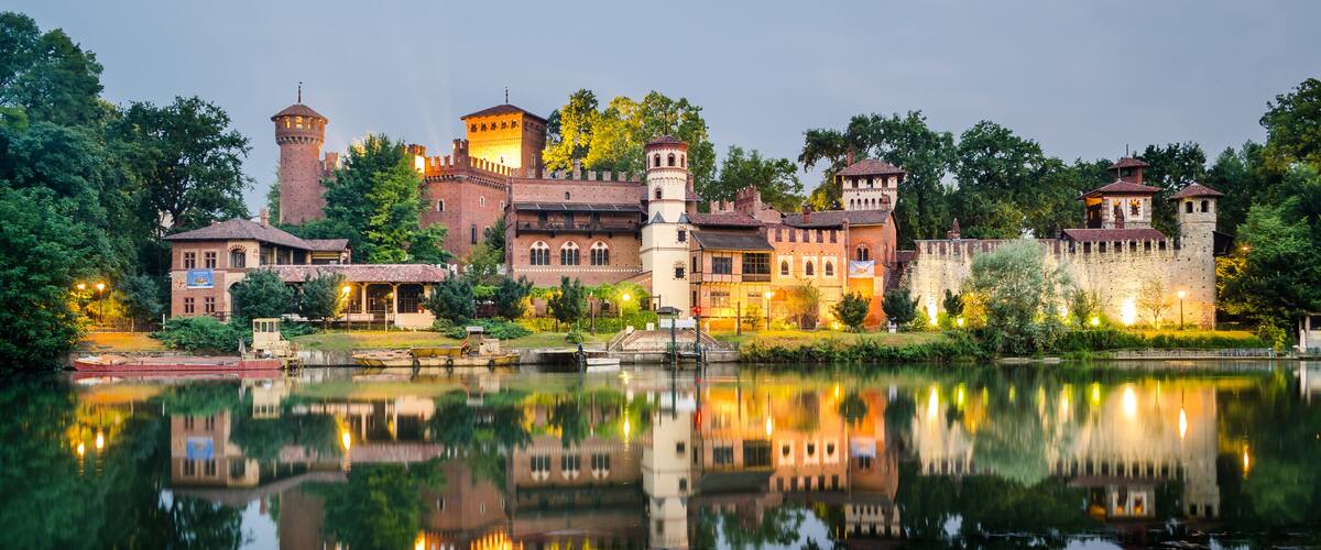 A historic castle with a moat, reflecting buildings, and lush greenery.