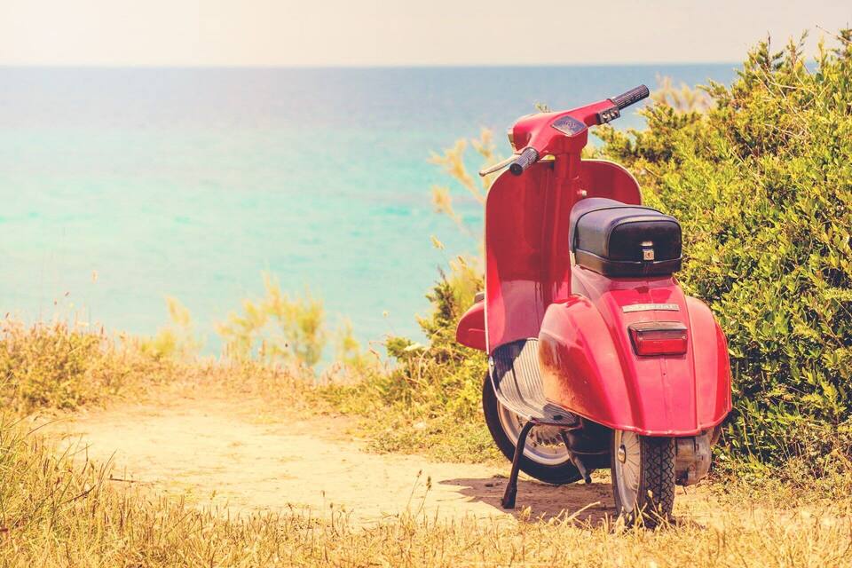 A red scooter parked on a dirt path with a view of the ocean and greenery.