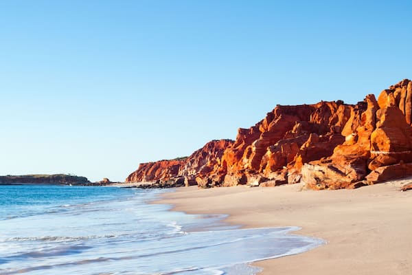 A beach with clear blue water, sandy shore, and red rock cliffs.