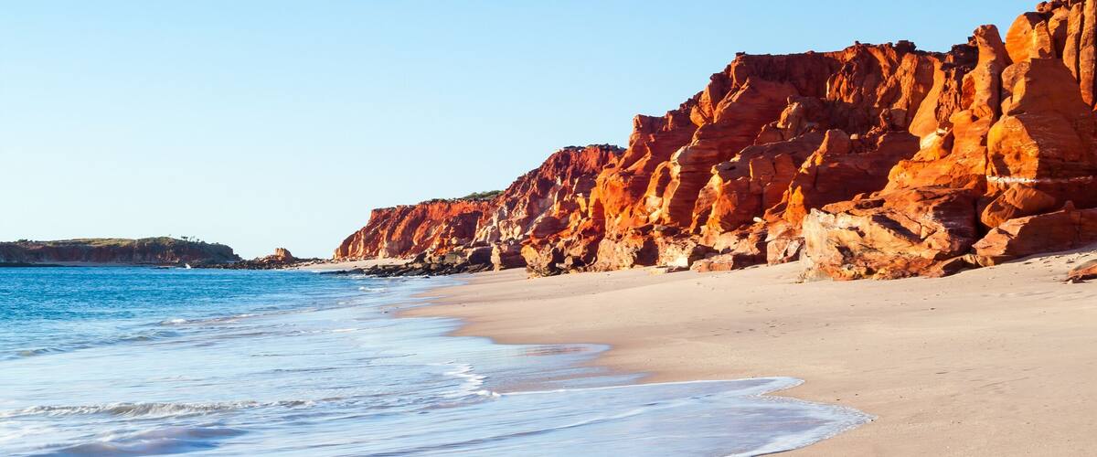 A beach with clear blue water, sandy shore, and red rock cliffs.