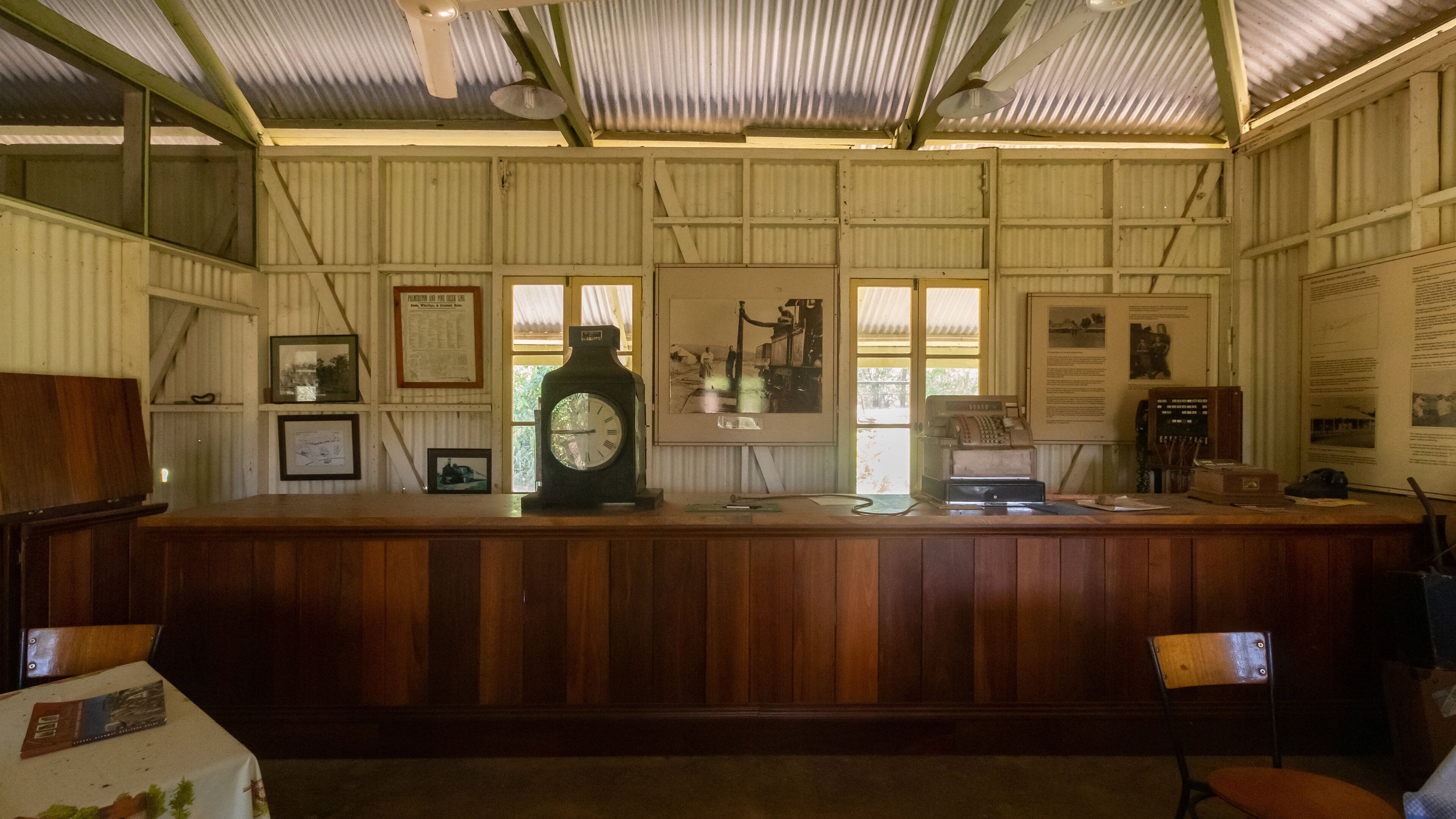 A wooden counter with a large clock, framed pictures, and vintage telephones in a rustic room with corrugated metal walls and a sloped ceiling.