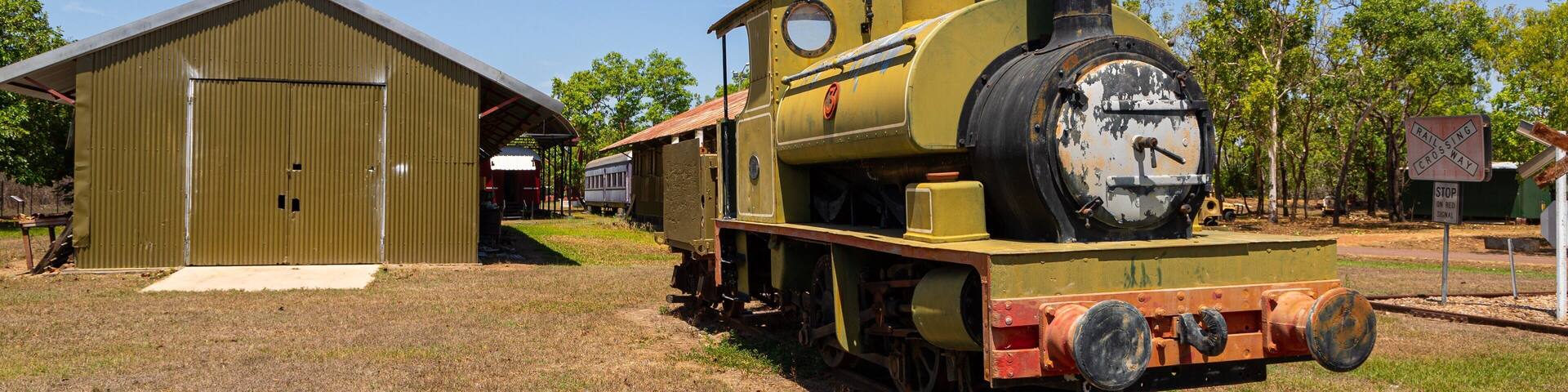An old yellow steam locomotive with a black smokestack, parked next to a green metal building.