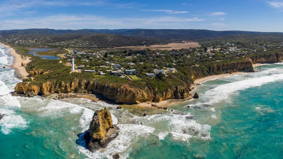 An aerial view of a coastal town with a lighthouse, clear blue waters, and a forested area.