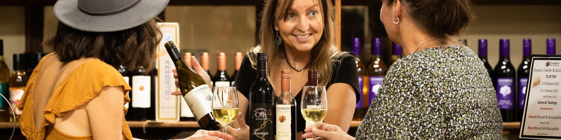 Três pessoas num bar, com garrafas de vinho ao fundo.
