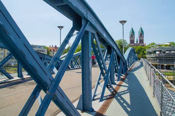 Eine blaue Stahlbrücke mit einem Fußweg und einem Radfahrer. Im Hintergrund sind zwei Gebäude mit begrünten Dächern und ein Gebäude mit rotem Dach zu sehen.