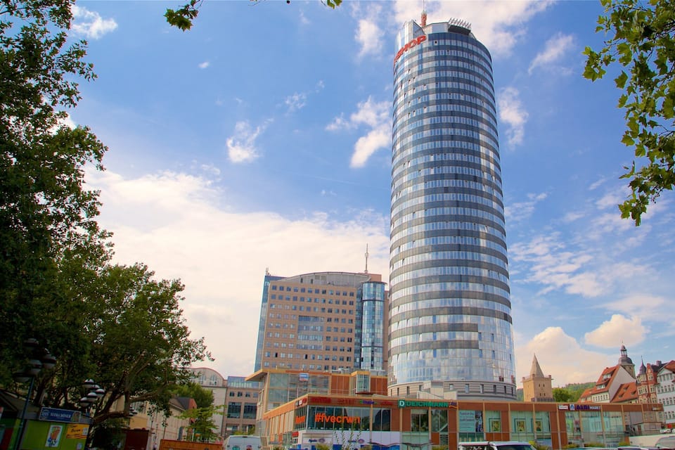 A modern cylindrical building with a glass facade, surrounded by traditional European architecture and trees.