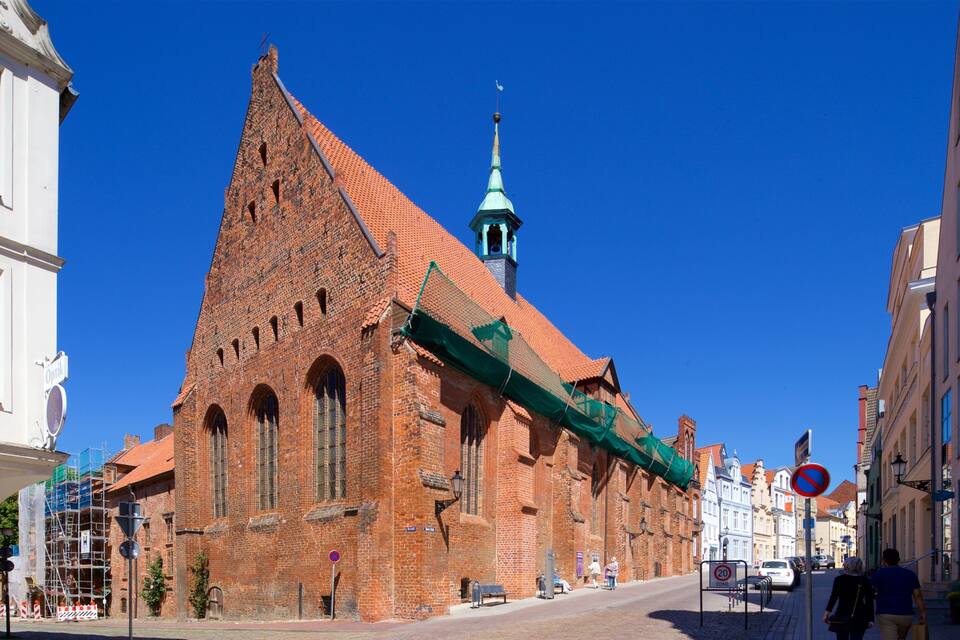 Eine historische Backsteinkirche mit einem grünen Turm, ein weißes Gebäude mit grünen Fenstern und ein Verbotsschild für Parkverbote an der Straße.