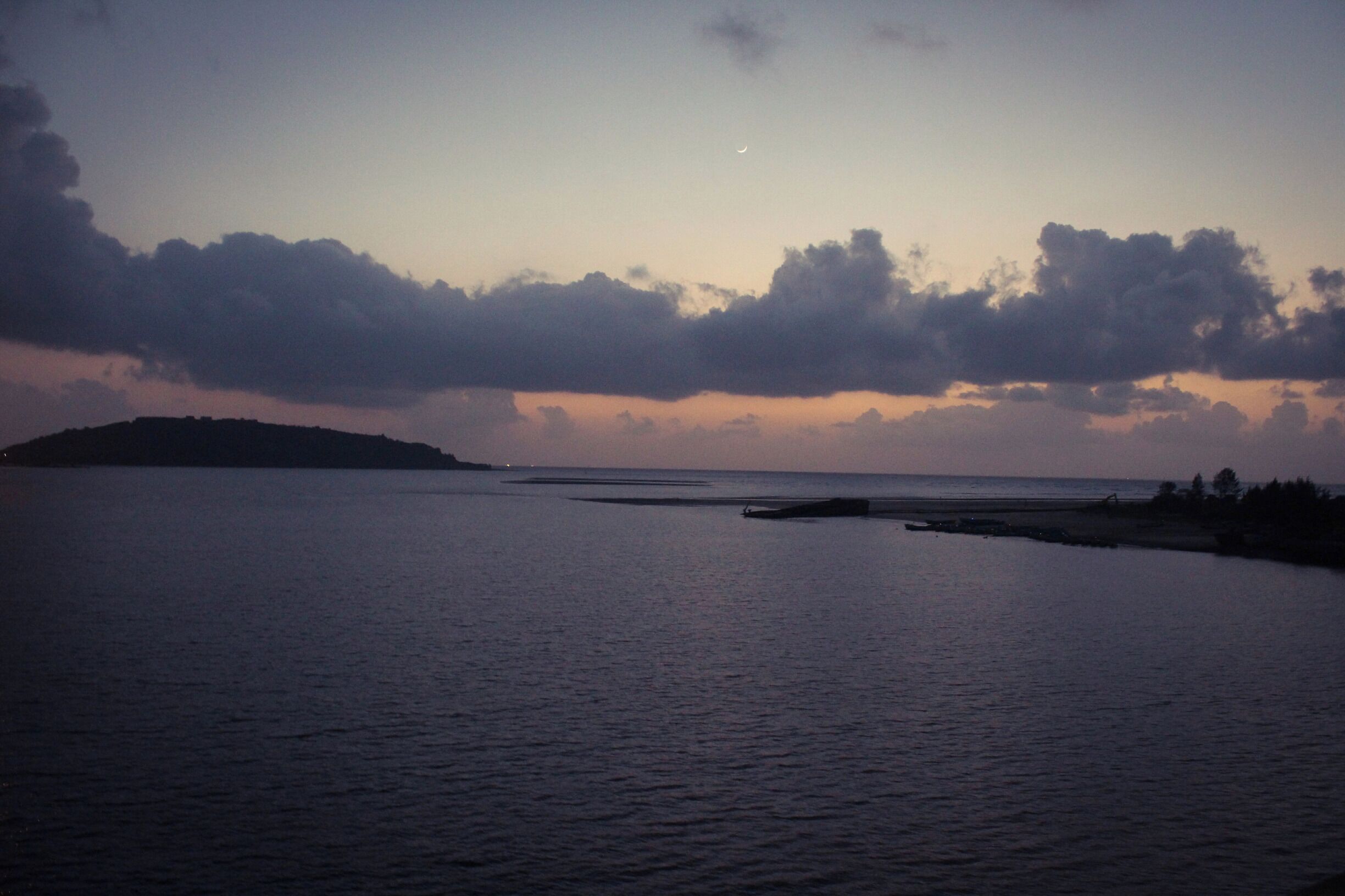 A calm sea at dusk with a crescent moon and clouds.