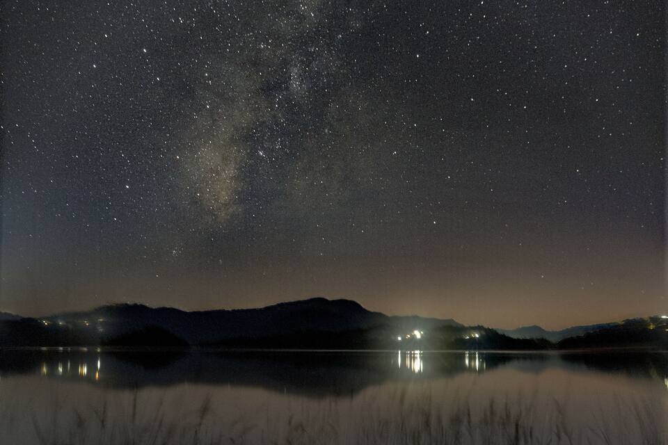 Un cielo notturno stellato sopra un lago tranquillo, con montagne in lontananza e alcune luci all'orizzonte.