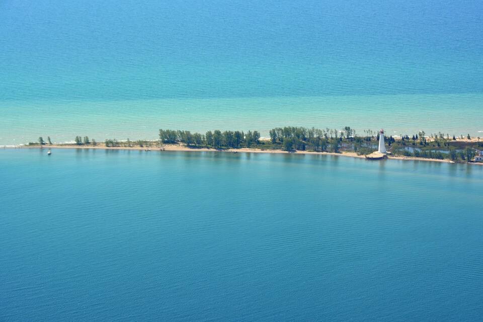 Paisaje costero con un faro, mar tranquilo y una orilla bordeada de árboles.
