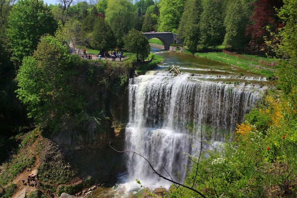 A waterfall cascading over a rocky cliff, surrounded by lush greenery and a stone bridge in the background.