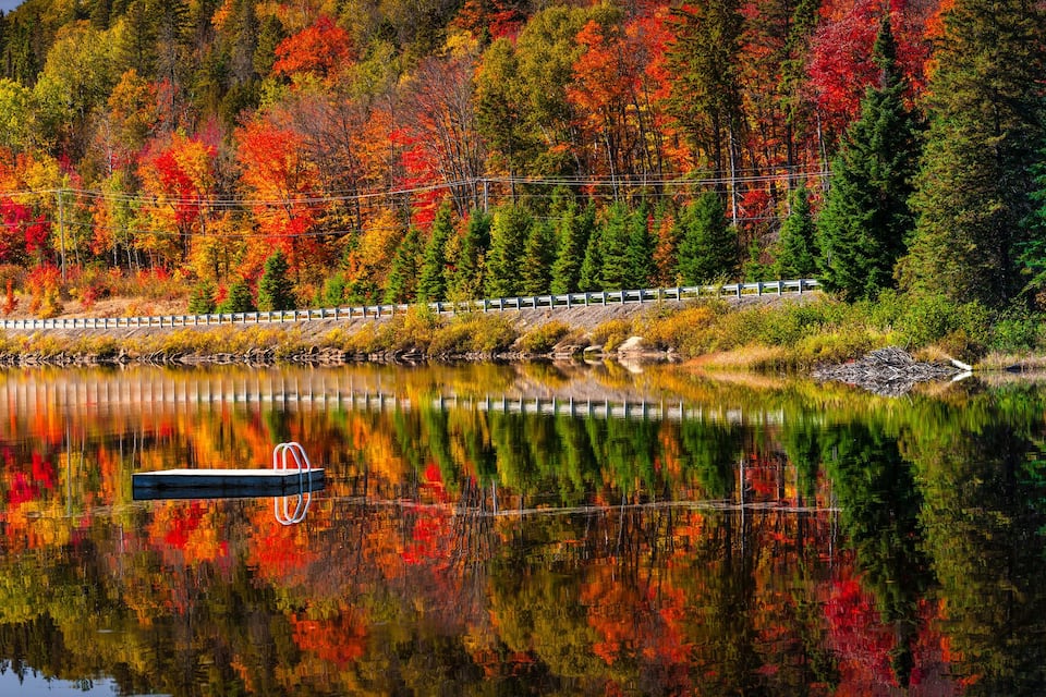 A lake reflecting colorful autumn trees, a train on tracks, and a dock.