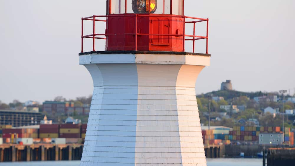 A white and red lighthouse with a clock, surrounded by a fenced area and a body of water.