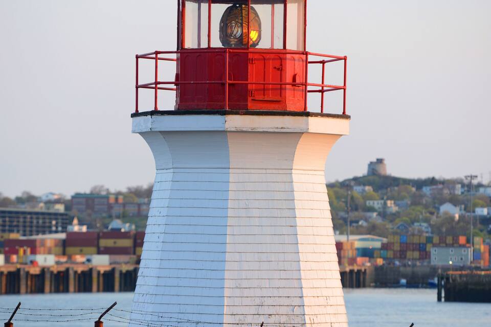 Un faro blanco y rojo con un reloj, rodeado por una zona cercada y una extensión de agua.