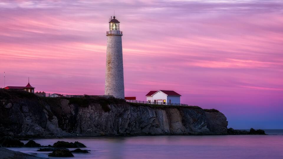 A lighthouse on a rocky cliff with a pink sky at dusk.
