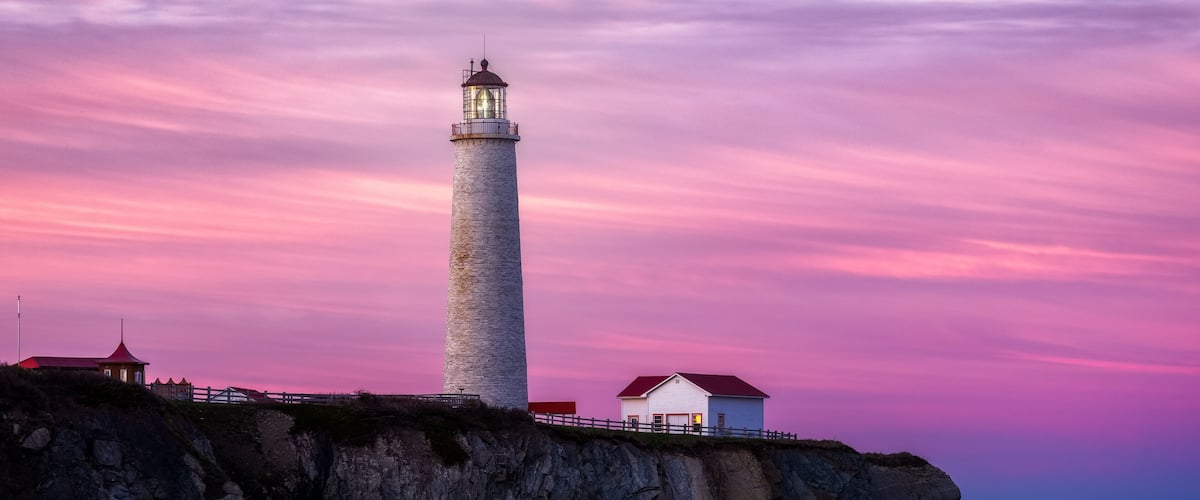 A lighthouse on a rocky cliff with a pink sky at dusk.