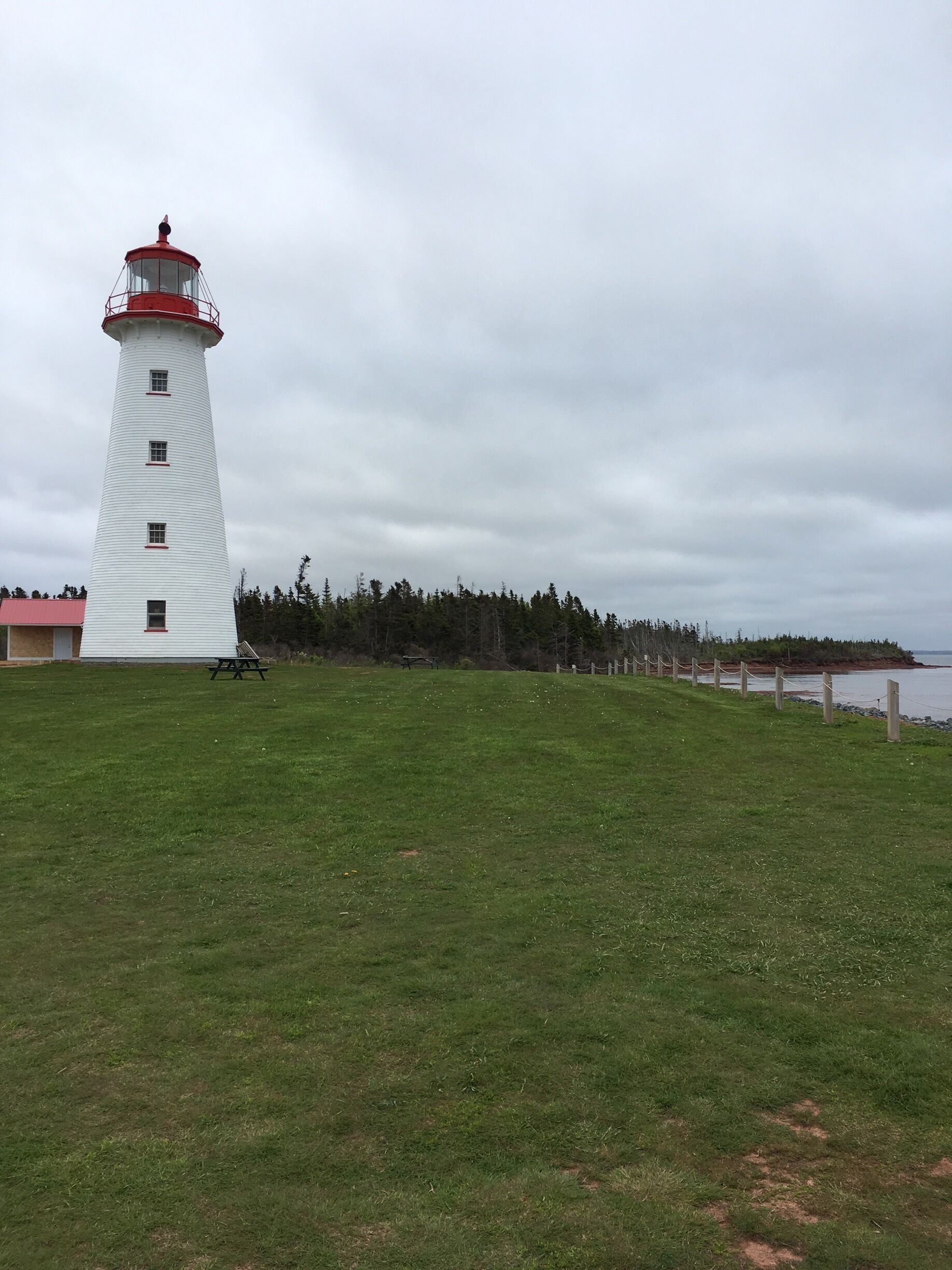 A white lighthouse with a red top, a grassy field, a wooden fence, and a body of water in the background.
